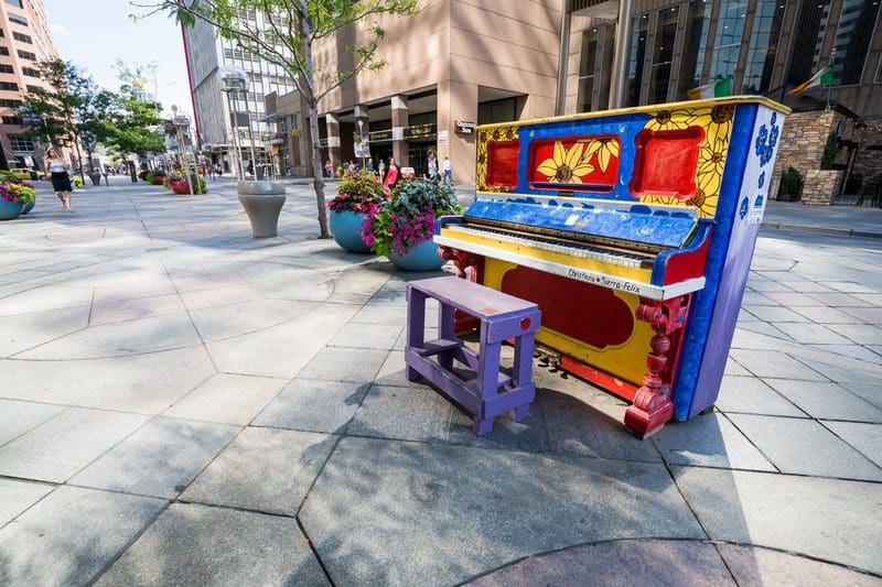 Colorful painted piano on a pedestrian walkway in downtown Denver, with nearby buildings, planters, and people walking in the background.